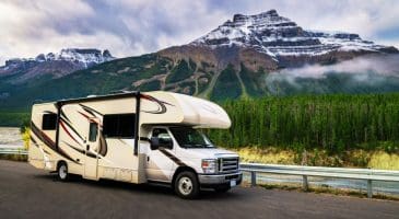 Motorhome parked on a scenic mountain highway with snow-capped peaks, evergreen forest, and low clouds in the background.
