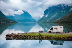 White Class C motorhome parked beside a mountain lake with snow-capped peaks reflected in the water